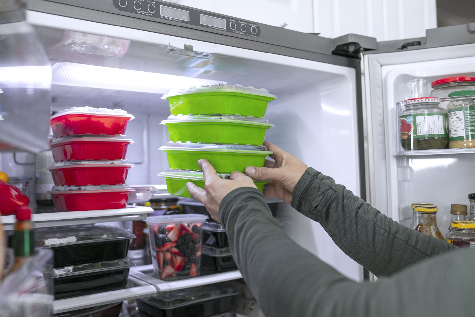 A person places stacked plastic food containers filled with leftovers inside a refrigerator with various jars, bottles, and food items.